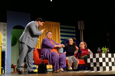 Das Bild zeigt eine emotionale Szene auf einer Theater-Bühne. Vier Personen streiten miteinander. Man sieht die bunte Kulisse mit einer blauen Wand, einem gelben Vorhang und einer gestreiften Lampe