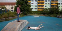 blaufarbige Skaterbahn mit einer auf dem Bauch liegenden Person und einer auf einer Rampe stehenden Person, die auf die Liegende sieht.